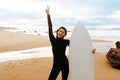 Young fit woman standing with surfboard on beach by seaside, smiling at camera and gesturing peace sign Royalty Free Stock Photo