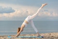 Young fit woman in white sportswear practices yoga asana pose on the beach on bright sunny day. Downward facing dog with one leg Royalty Free Stock Photo