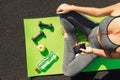 Young fitness sportswoman sitting and using smartphone in gym. Girl on yoga mat with botte of water, dumbbell and ealthy apple