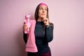 Young fitness woman wearing sport clothes and towel drinking water from take away bottle serious face thinking about question, Royalty Free Stock Photo