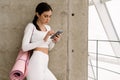 Young focus woman using mobile phone while standing with yoga mat