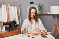 Young focused concentrated woman sewer sitting at her workplace in atelier near sewing machine with scissors in hands, thinking Royalty Free Stock Photo