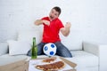 Young football fan man watching game on tv in team jersey celebrating goal crazy happy on couch