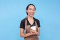 A young and friendly asian female barista chuckling while holding an order of coffee. Wearing a brown apron and black shirt and