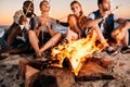 young friends roasting marshmallows at bonfire while sitting at sandy beach