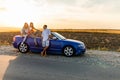 Young three friends standing near convertible car and talking each other have fun on beauty sunset on the road Royalty Free Stock Photo