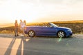 Young three friends standing near convertible car and talking each other have fun on beauty sunset on the road Royalty Free Stock Photo