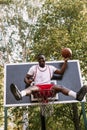 Young funny African American basketball player in a white T-shirt and red shorts sits in a basket on the basketball