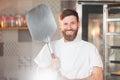 A young funny baker with a pizza spatula in his hands against the backdrop of an oven.