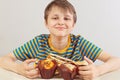 Young funny boy in a striped shirt at the table with muffins and sandwich on white background