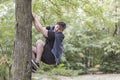 Young funny caucasian man climbs up to the tree with fair or horror of something below. White wireless earphones, casual sportswea