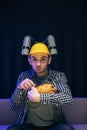 Young funny man with beer helmet on the head eating chips while watching movie on the sofa in the living room Royalty Free Stock Photo