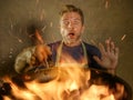 Young funny and messy home cook man with apron in shock holding pan in fire burning the food in kitchen disaster and domestic cook