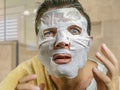 Young funny and messy man applying beauty facial mask looking to the mirror surprised and horrified in skin care aging concept and