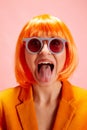Young funny woman in bright orange wig and retro sunglasses looking at camera opening her mouth and shows tongue. Royalty Free Stock Photo