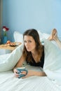 Young funny woman having a breakfast on tray on the bed with cup of coffee under coverlet Royalty Free Stock Photo