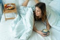 Young funny woman having a breakfast on tray on the bed with cup of coffee under coverlet Royalty Free Stock Photo