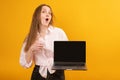 Young funny woman holds laptop with empty black screen workspace area show like gesture. Yellow background. Copy space, mock up Royalty Free Stock Photo