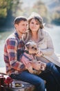 Young future parents and their dog in a funny costume sitting on a wooden bridge and having picnic near lake Royalty Free Stock Photo