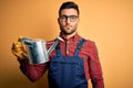 Young gardener man wearing working apron and gloves holding watering can with a confident expression on smart face thinking