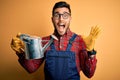 Young gardener man wearing working apron and gloves holding watering can very happy and excited, winner expression celebrating Royalty Free Stock Photo