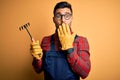 Young gardener man wearing working apron using gloves and tool over yellow background cover mouth with hand shocked with shame for