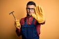 Young gardener man wearing working apron using gloves and tool over yellow background with open hand doing stop sign with serious Royalty Free Stock Photo