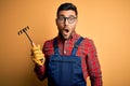 Young gardener man wearing working apron using gloves and tool over yellow background scared in shock with a surprise face, afraid