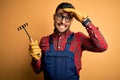 Young gardener man wearing working apron using gloves and tool over yellow background stressed with hand on head, shocked with