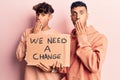 Young gay couple holding we need a change banner covering mouth with hand, shocked and afraid for mistake