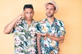 Young gay couple of two men wearing summer hat and hawaiian shirt confused and annoyed with open palm showing copy space and