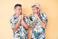 Young gay couple of two men wearing summer hat and hawaiian shirt laughing nervous and excited with hands on chin looking to the Royalty Free Stock Photo