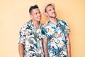 Young gay couple of two men wearing summer hat and hawaiian shirt looking away to side with smile on face, natural expression Royalty Free Stock Photo