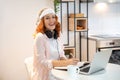 Young ginger smiling woman taking notes and working on laptop at home. Wearing christmas hat on head and headset on neck Royalty Free Stock Photo