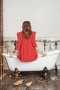 Young ginger woman sit on the edge of antique bathtube on lion paws, back to the camera, worn bright red dress.