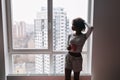 Young girl, african teen looking through big window with high-rise buildings and holding coffee, tea mug in room at home Royalty Free Stock Photo