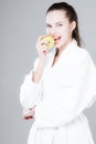 A young girl bites an apple, stands in a white robe on a gray background. Dental health and proper nutrition concept