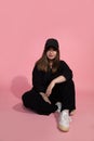 Young girl in black baseball cap sits in studio on the floor on pink background looks into the frame