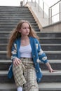 A young girl, blonde with long hair, in a blue cardigan is sitting on the steps of a gray marble staircase Royalty Free Stock Photo