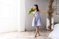 A young girl in a blue dress stands by the bed in a spacious white room and inhales the scent of a large bouquet of Royalty Free Stock Photo