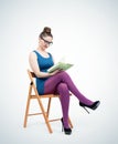 Young girl in a blue mini dress with glasses sits on a chair and reads a book Royalty Free Stock Photo