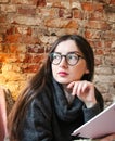 A young girl with dark long hair and glasses sits on a sofa in a cafe against a brick wall. Cute young woman holds a notebook in Royalty Free Stock Photo