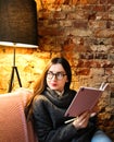 A young girl with dark long hair and glasses sits on a sofa in a cafe against a brick wall. Cute young woman holds a notebook in Royalty Free Stock Photo