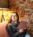 A young girl with dark long hair and glasses sits on a sofa in a cafe against a brick wall. Cute young woman holds a notebook in Royalty Free Stock Photo