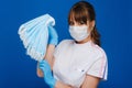 A young girl doctor in a medical mask holds a lot of masks in her hands. A nurse poses on a blue background