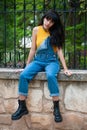 Young girl dressed in yellow and wearing a denim jumpsuit sitting on a stone wall in a park with autumn green and yellow colors Royalty Free Stock Photo