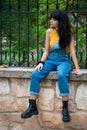 Young girl dressed in yellow and wearing a denim jumpsuit sitting on a stone wall in a park with autumn green and yellow colors Royalty Free Stock Photo