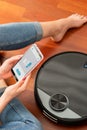 Young girl feet at home using a smart phone to control a round smart automatic vacuum, machine housework robot cleaning the floor