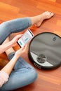 Young girl feet at home using a smart phone to control a round smart automatic vacuum, machine housework robot cleaning the floor