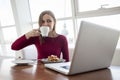 Young girl freelancer sits in a cafe with a laptop, drinks coffee and has breakfast, a student eats in the morning with a computer Royalty Free Stock Photo